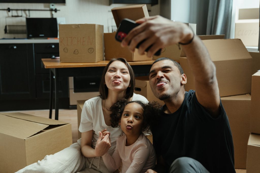 Family taking selfie in front of moving boxes, making silly faces and looking happy.